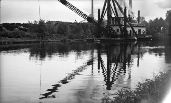 View across water of the dredge "Winneconne" on the Fox River canal. Although undated the photograph was probably taken during the 1940s.  The "Winneconne" was the second steel self-unloading dredge built by the Leathem Smith Company for the Army Corps of Engineers.