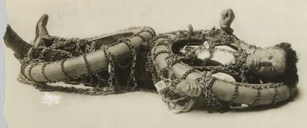 Harry Houdini, the well-known escape artist, was born in Budapest, Hungary and raised in Appleton, Wisconsin. At the time of his immigration his name was spelled Ehrich Weisz. He is lying down wrapped in chains with heavy locks, and what appear to be two automobile tires.
