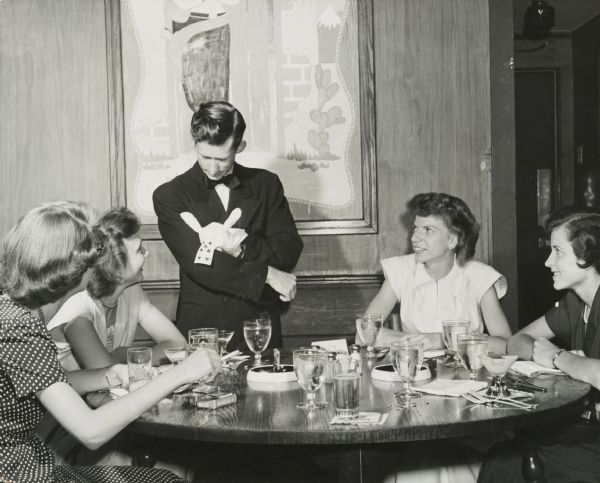 Magician Eddie Tullock (1919-2007) performing at a restaurant. Minnesota-born Tullock was the originator of "trade show magic," the concept of presenting a commercial message to a small audience by virtue of his personality and skill.