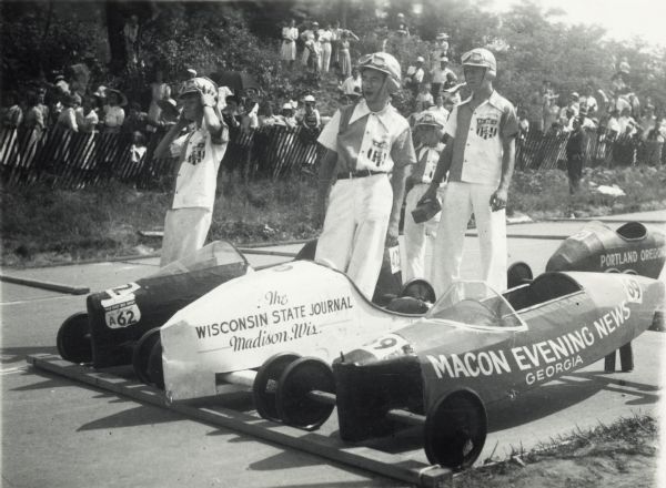 Thirteen year-old James Borden (standing in the middle) of Madison, Wisconsin, the 1940 Madison, Wisconsin soap box derby champion, waiting for his turn to compete at the All-American Soap Box Derby, Derby Downs, Akron, Ohio. He is standing next to his Wisconsin State Journal-sponsored car. James did not win his heat.