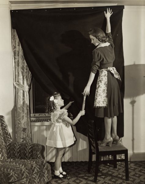 Publicity photograph of a woman hanging blackout curtains, with the assistance of her young daughter. The photograph was taken to provide illustration for a public relations campaign created by the Advertising Women of New York and chaired by Dorothy Dignam to support the war effort.