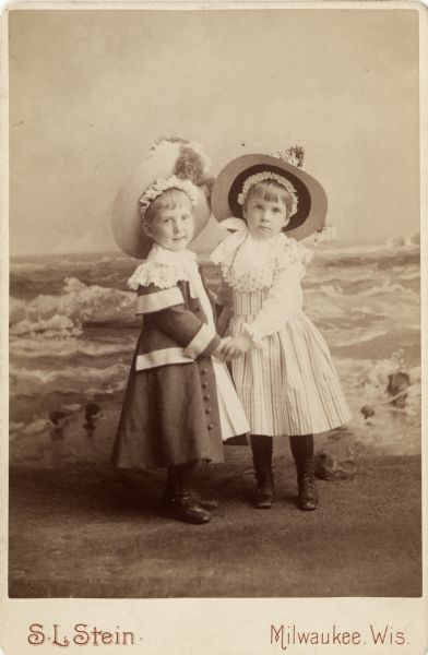 Full-length cabinet card of two young children in front of a painted backdrop of the ocean. They are attired in coats, dresses and hats, and are holding hands. 

The children are at right Hilda Johnston (1883-1974), age 4, and at left John "Jack" Thorsen Johnston (1884-1939), age 3, the children of John and Ethelinda Johnston (1856-1947) of Milwaukee.  John Johnston (1836-1904) was a successful Milwaukee banker.

The hats and Jack's coat were made by Mrs. Emmons.