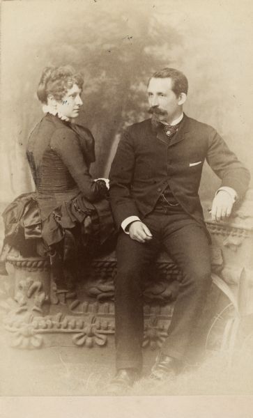 Cabinet card of John (1836-1904) and Ethelinda (1856-1947) Johnston sitting on a decorative wall in front of a painted backdrop of trees.  John, a successful Milwaukee banker, married his second wife Ethelinda Thorsen on September 1, 1881.