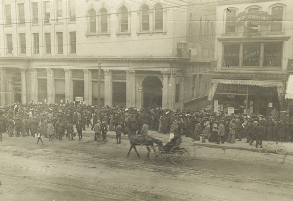 Elevated view of street scene of a run on the First National Bank. The sidewalk is crowded with people, spilling into the street. A horse-drawn buggy is driving past in the foreground. A tailor and a music store are on the right.

On Saturday, April 22, 1905 the trustees of the First National Bank discovered that the bank's president Frank Bigelow had embezzled $1,500,000. He was arrested on Monday, April 24th and was sentenced to ten years hard labor on June 15th or 16th, 1905. The trustees were able replace the funds fairly quickly, so this run probably occurred the week of April 24th.