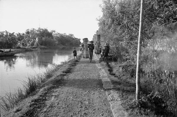 Canal in a hamlet near Phat Diem, described by photographer David Schoenbrun as part of an extensive system of irrigation. On a path along the canal a man is carrying rugs hanging from a wooden yolk over his shoulders. Children and other people on bikes are with him. On the far left are people with a boat.