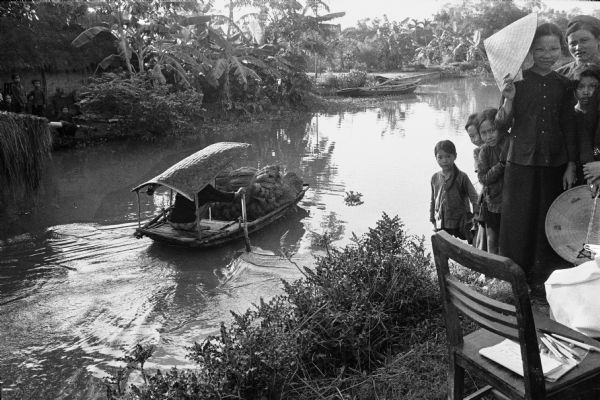 Person steering a sampan, loaded with what appears to be fiber, on an irrigation canal near the North Vietnamese city of Phat Diem. On the right is a group of children and adults. Brushes and papers lie on a chair in the right foreground. Another group of children is on the far bank, in front of a thatched roof building.