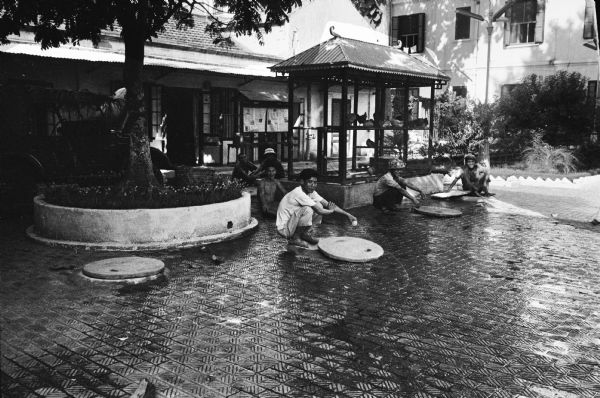 Individual air raid shelters in the courtyard of the hotel where journalist David Schoenbrun was staying in Hanoi in 1967. There is a chicken coop behind the group of men squatting near the air raid shelter entrances which are holes with covers in the brick courtyard.