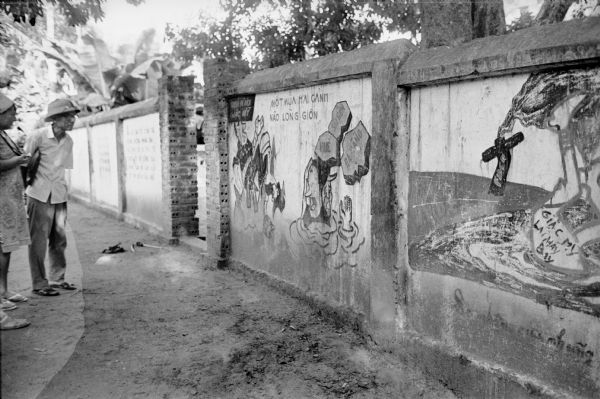 Dorothy Schoenbrun (far left) views an anti-American wall mural in the North Vietnamese village of Phu Xa. A man is standing next to her. Schoenbrun was traveling with her husband, David Schoenbrun, an American journalist.