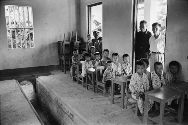 Kindergarten classroom in the North Vietnamese village of Phu Xa, showing the ditch in the center of the room that ran to the air raid shelter. Two men stand in an open doorway. This village was visited by American journalist David Schoenbrun.