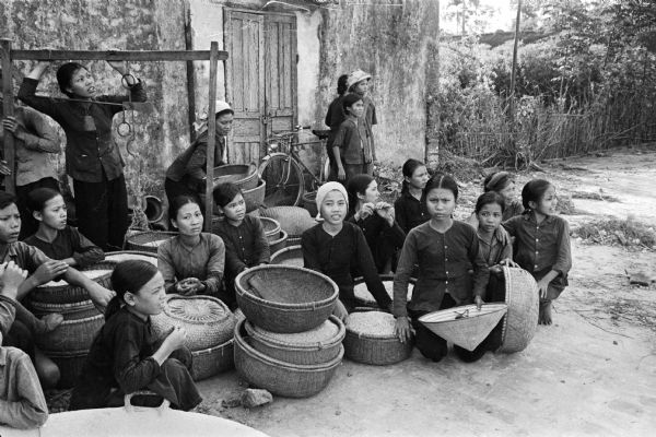 A group of people sit and stand outdoors with a group of baskets at a collective farm near Phat Diem, North Vietnam, visited by American journalist David Schoenbrun. Schoenbrun indicated that American airplanes were passing overhead while he took these pictures.