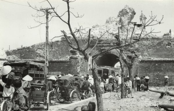 A crowd of pedestrians, trucks, and cyclists on a road waiting to go through an arch in a damaged building amid the post-Tet destruction in Hue.