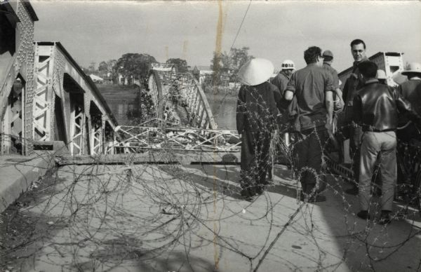 Journalist Robert Shaplen, facing the camera, at a bombed bridge over the Perfume River at Hue, where crowds of pedestrians and cyclists were attempting to cross. Shaplen, whose papers are part of the Archives of the Wisconsin Historical Society, was well known for his coverage of the war in Vietnam for "The New Yorker" magazine. There are coils of barbed wire in the foreground.