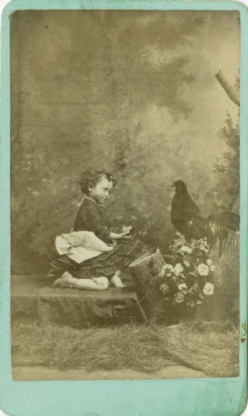 Carte-de-visite of a young girl posed with a rooster in front of a painted backdrop. She wears a dress and holds a partially eaten apple in her hand. She and the rooster are gazing at one another. The rooster is perched on a tree stump decorated with flowers.