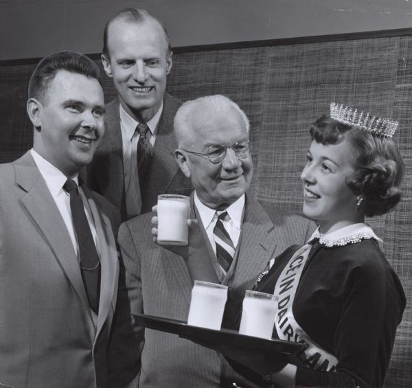 The Alice in Dairyland holding a tray of glasses of milk, posing with three men.