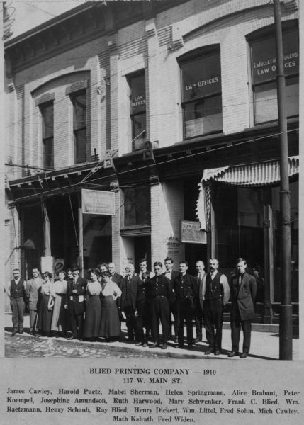 The staff of Blied Printing Company posed in front of the building at 117 W. Main Street. Employees listed are James Cawley, Harold Puetz, Mabel Sherman, Helen Springman, Alice Brabant, Peter Koempel, Josephine Amundson, Ruth Harwood, Mary Schwenker, Frank C. Blied, Wm. Raetzmann, Henry Schaub, Ray Blied, Henry Dickert, Wm. Littel, Fred Sohm, Mich Cawley, Math Kalrath, Fred Widen.