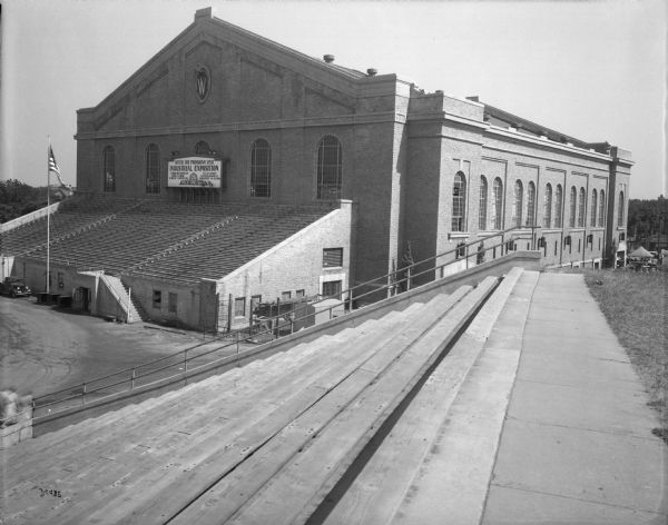 Exterior view of the back of the University of Wisconsin-Madison Field House. A sign reads "After the Program Visit Industrial Exposition." The Wisconsin Centennial Industrial Exposition was held in the Field House in June and July.