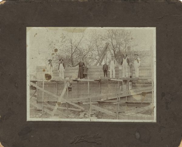 Construction crew and observers standing on a scaffold. Trees, a wooden tripod with a block and tackle and the roof of a house are visible in the background. On the left is Henry Mathews, early African-American resident of Fox Lake. He was a stone mason for many years.