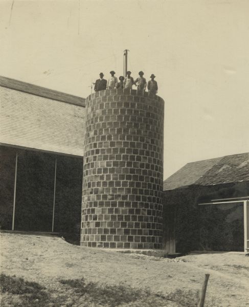 Construction crew on Kastenmeier farm. They are standing at the top of a silo they are building. A barn is on the left and a shed is on the right. Names from left to right: Ira Schroder, Henry Mathews (in his 70s), Sam Mathews (oldest son of Henry), Fred Koufel, Clifford Mathews (son of Henry), Ollie Kastenmeier.