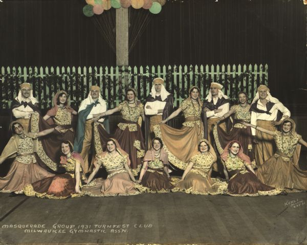 Masquerade Group, Turnfest Club, Milwaukee Gymnastic Association. Five men and eleven women are dressed up in masquerade clothing, and all are wearing headcloths. An ivy covered picket fence appears in the background. The photograph has been colorized.