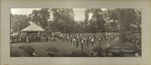 Children’s class of the Milwaukee Turners, gathered outside in a park. George Brosius appears on the platform and several people are wearing band uniforms. Behind the platform is a small building labeled "Tintypes." It may be drizzling since the spectators are holding umbrellas.
