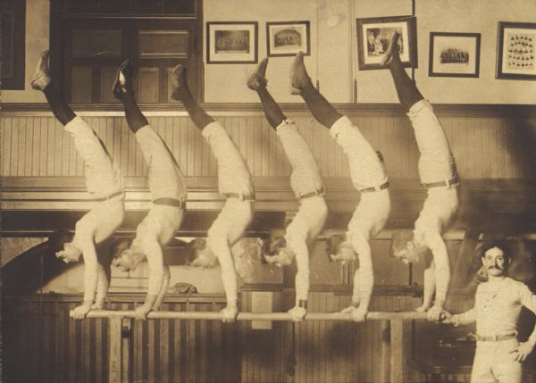 Six men from Milwaukee Turners doing handstands on parallel bars in a gymnasium. A seventh man stands to the right. All are wearing uniforms.