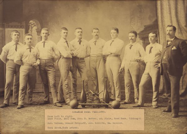 Adult men, dressed in uniforms, posing in front of a painted backdrop with sporting equipment indoors at the Appleton Turners Festival. Their teacher stands on the right, dressed in a suit.