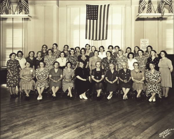 The Ladies Auxiliary of the Milwaukee Turners posing indoors. They are wearing dresses. An American flag hangs behind them.


