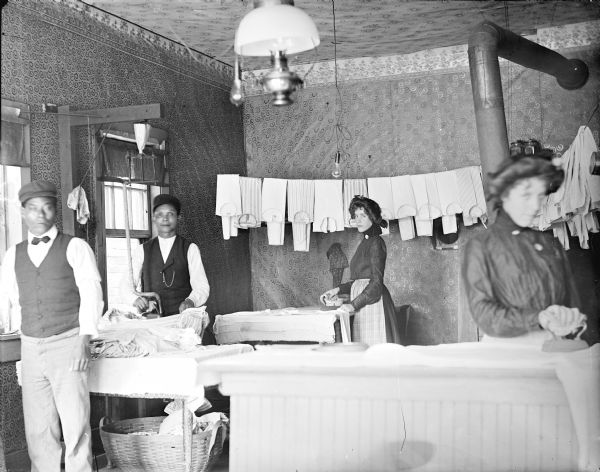 Interior view of Yep Sing's Chinese Laundry. Clothing is hanging on a line and Yep Sing (second from left), Anna Erickson (center), and Carrie Aldra (right) are pressing clothes with hot irons. A man in a vest, hat and bow tie is standing on the left.