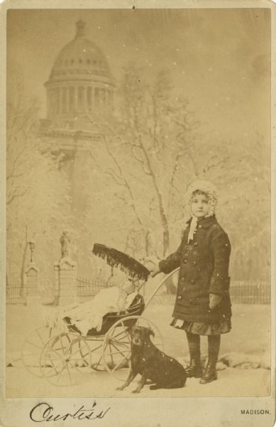 Cabinet card studio portrait of a young girl, posed with a doll in a baby carriage and her dog. The background is a painted backdrop of the Wisconsin State Capitol building in winter. She is wearing a dress, stockings, shoes, coat, mittens and a white fluffy hood. Simulated snow was added to the negative before printing.