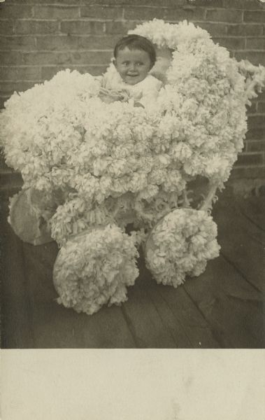 Photographic postcard of a young, smiling child sitting in a baby carriage covered in flowers.