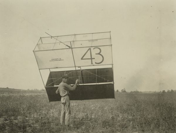 A man in a field holds a very large kite just prior to launching it. On the kite is painted the number 43 and "Property of the U.S. Weather Bureau." He is wearing work clothes and a cap.