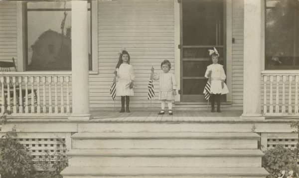 Photographic postcard of three young children standing on a porch, each holding an American Flag. They are wearing dresses, stockings and shoes. Two are girls wearing bows in their hair.