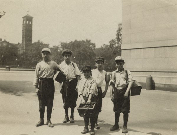 Five boys stand on the sidewalk holding their shoe shining tools and supplies. They are wearing work clothes; shirts, knickers, stockings, shoes and caps. Two boys wear Grafin Shoe Polish caps. A church tower is visible in the background.