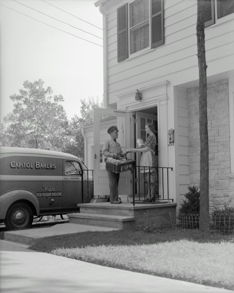 A woman standing on her doorstep buys bread from a deliveryman. His truck is parked in the driveway. Text on the truck reads "Capitol Bakers, 'Right' to your door." She is wearing a dress with an apron and he is wearing a uniform, bow tie and a cap.