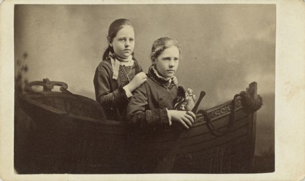 Carte-de-visite of a studio portrait of two girls posed in a small "boat" in front of a painted backdrop. They are wearing fancy dresses, and one girl holds an oar. On the back is the note, "Dolly Lawrence, 13 yrs. 1878."