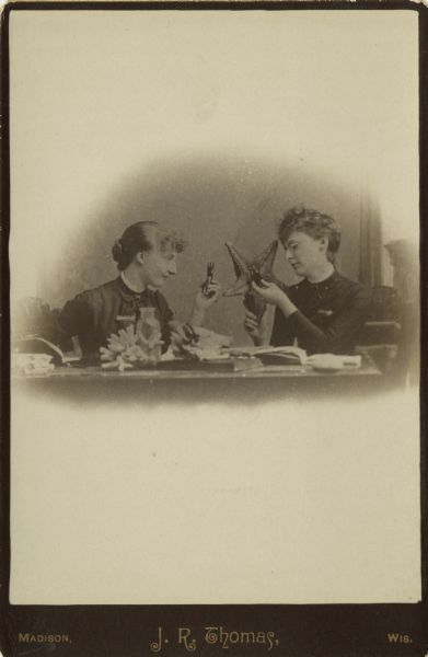 Cabinet card of two women sitting at a table examining a starfish and other underwater specimens using magnifiers. Open books appear on the table, probably for reference. They are wearing dresses with pins at the collar.