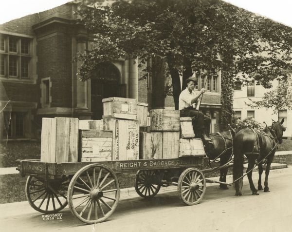 C.W. Jarvis' horse-drawn delivery wagon loaded with crates in front of 201 North Carroll Street, the location of the old Madison Public Library. He is sitting on the wagon seat, holding a buggy whip, gazing at the camera, and wearing work clothes and a hat. Painted on the wagon are the words: "Trunks," "Freight & Baggage" and "C.W. Jarvis." Some of the crates have labels, "Wisconsin Historical," "Library, Madison, Wis.," "Smith Premier Typewriter" and "United States Army, Q.M.C. Supplies, General, Must Not Be Delayed."
