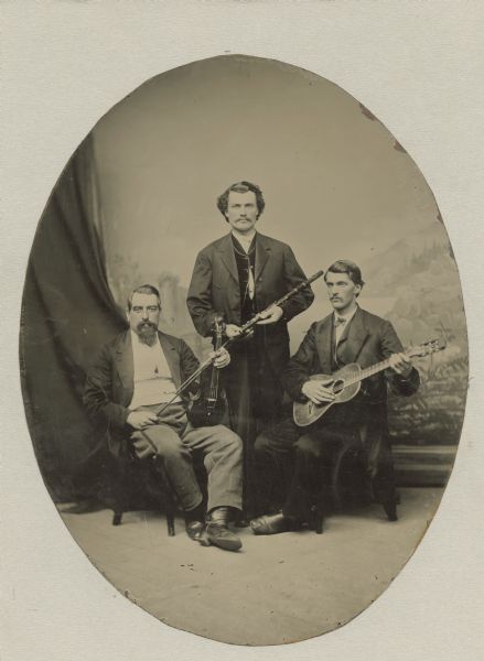 Ferrotype studio portrait of a musical trio in front of a painted backdrop. One man on the right sits holding a guitar, a man stands in the middle holding a flute, and the man on the left sits holding a violin. The photograph was cropped in the shape of an oval and mounted on a piece of mat board. A typewritten label attached below reads, "Musical group, probably in Milwaukee, Wis., about 1875(?). One of the seated men, (probably the violinist at left) was called 'Papa Hess'. Standing is Anton Kayser who came with his family as a small boy to Sauk City in 1858. At 17, he ran away to enlist and was a Civil War drummer boy and later a cavalryman and eventually "head" of the Grand Army of the Republic organization in Milwaukee with his wife and five children and died there about 1922."