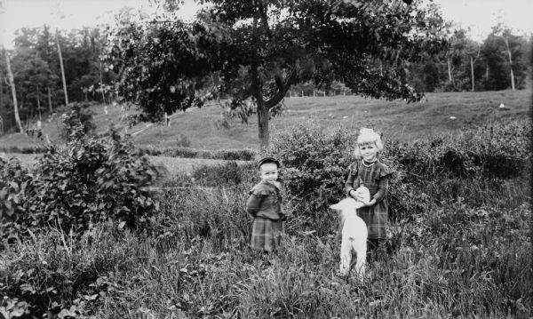 Glendora and Russell Watt, the children of Ephraim and Adelaide Watt are standing with a lamb in an open field. Glendora is holding a bottle for feeding the lamb. Both children wear plaid clothing and Glendora has a necklace.