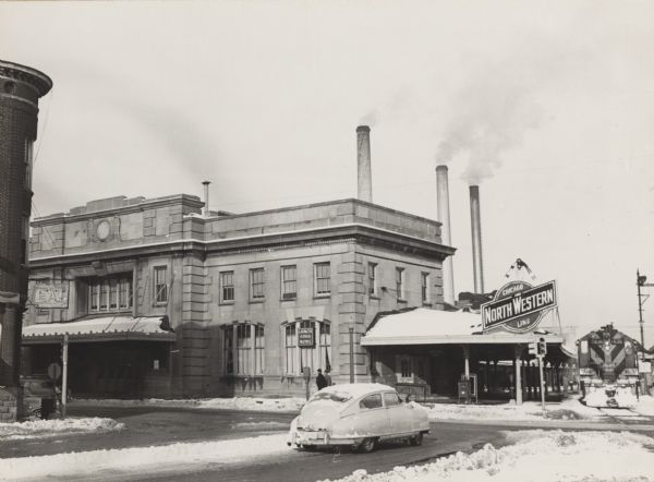 View across intersection of the Chicago and Northwestern railroad station at Blair and Wilson Streets. Three tall chimneys from the MG&E plant can be seen in the background.