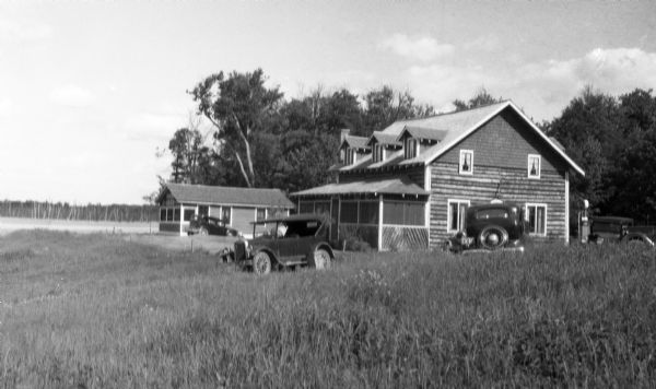 Old cars parked in tall grass next to a house with a porch; a gas pump is on the right. There is a smaller dwelling with a car parked next to it on the left, which appears to be on the shoreline of a lake.