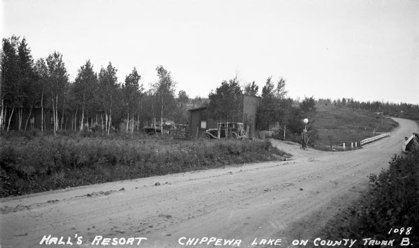 View across road of an automobile parked next to a building that has a gasoline pump in front of it by the dirt road of County Trunk B. There is a hand-painted sign pointing to "cabins". A cabin is on the left in a new growth of trees.