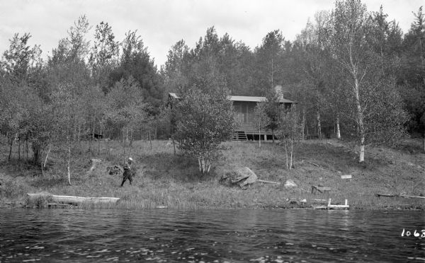 View from water of a man wearing a hat and boots walking along the shoreline. A cabin is located up on the bank.