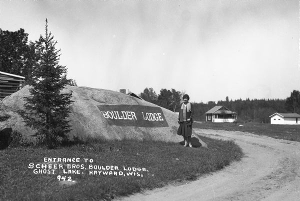A woman stands next to a boulder along a drive leading to cabins. "BOULDER LODGE" is printed on the boulder. Boulder Lodge is located on Ghost Lake.