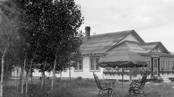 View from yard of a single story house with enclosed front porch. In the foreground is an outdoor umbrella over two chairs and a small table.