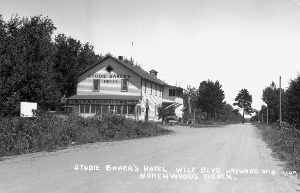 View from road of large two-story building situated along roadway. A gasoline pump is next to the building along with four fifty-five gallon drums positioned on their sides on a short base. There is a medium-sized pine tree growing in the middle of the road in the far background.
