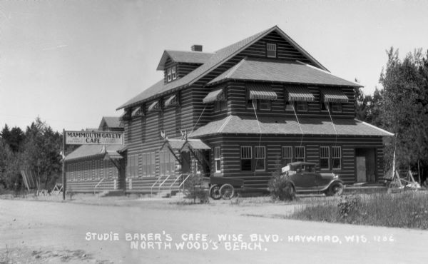 Large two-plus story log building with awnings over the windows, with a one-story attached cafe with a sign that says: MAMMOUTH GAYETY CAFE. There is a coupe-style car with a small trailer parked alongside the building.