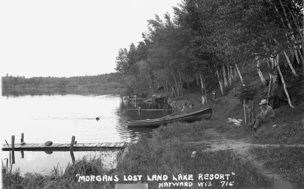View along shoreline of a man wearing a hat and sitting on a hillside. A dog retrieves a stick in the water. Wooden row boats are at docks in the background. Caption reads: "Morgan's Lost Land Lake Resort" Hayward Wis 715."