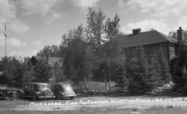 Two-story log building surrounded by immature trees on Moose Lake. There is a flagpole on the left, and automobiles are parked in the foreground.