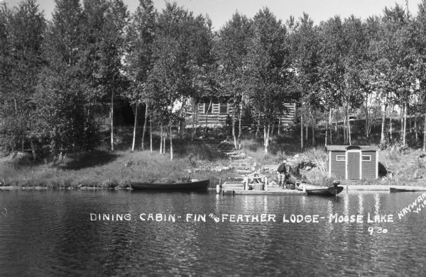 View over Moose Lake of log building on low hillside. A man and another person are standing on a dock next to a wood boat with a small motor attached, and another boat is tied along the shore.
